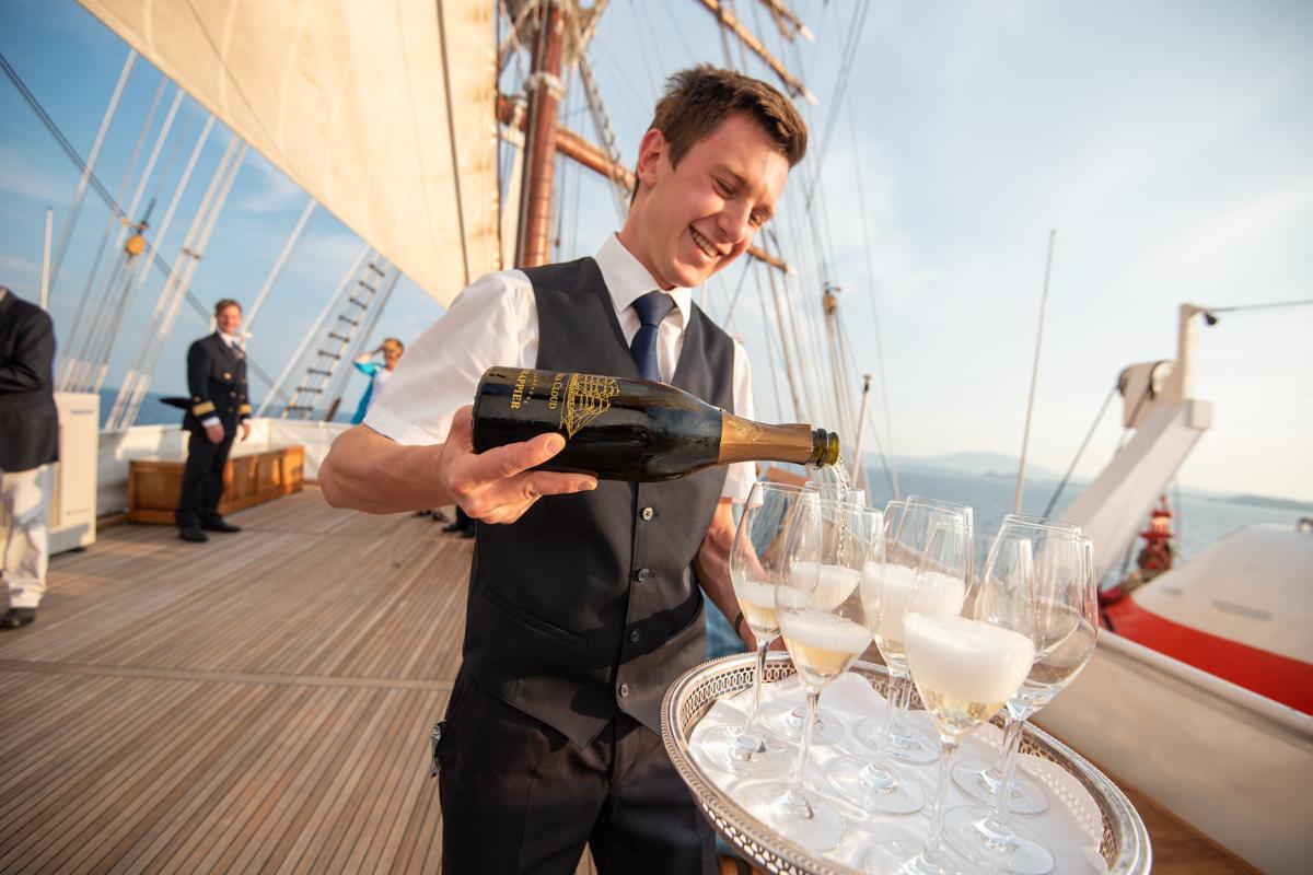 A Steward on the deck of the ship Sea Cloud pours champagne for guests.